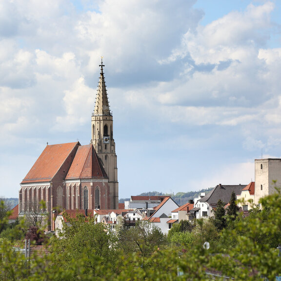 Eine malerische Aussicht auf eine Kirche mit einem hohen Turm und einem roten Ziegeldach, umgeben von malerischen Gebäuden und üppigem Grün unter einem bewölkten Himmel.