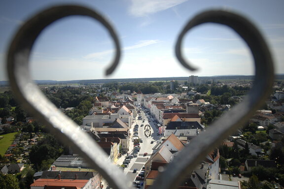 Blick auf die Stadt Neuötting mit einer von Gebäuden gesäumten Straße, gesehen durch ein herzförmiges Eisengeländer. Die Landschaft erstreckt sich unter einem blauen Himmel bis zum Horizont.